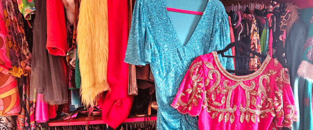Clothes hanging on racks in lots of bright colours. a bright pink and bright blue dress is hanging separately on display, sequined.