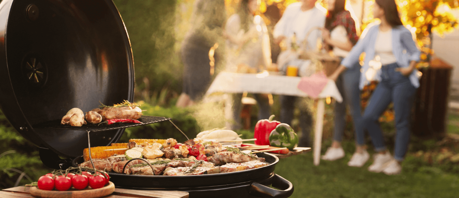 A barbecue in the foreground with food on it. In the background is blurred people standing around a table.
